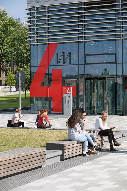 This picture shows building four of the new campus Derendorf of Hochschule Düsseldorf. The front of the building is of glass and steel, indoors you see red elements like the stairs and the walls. This building includes the lecture rooms.